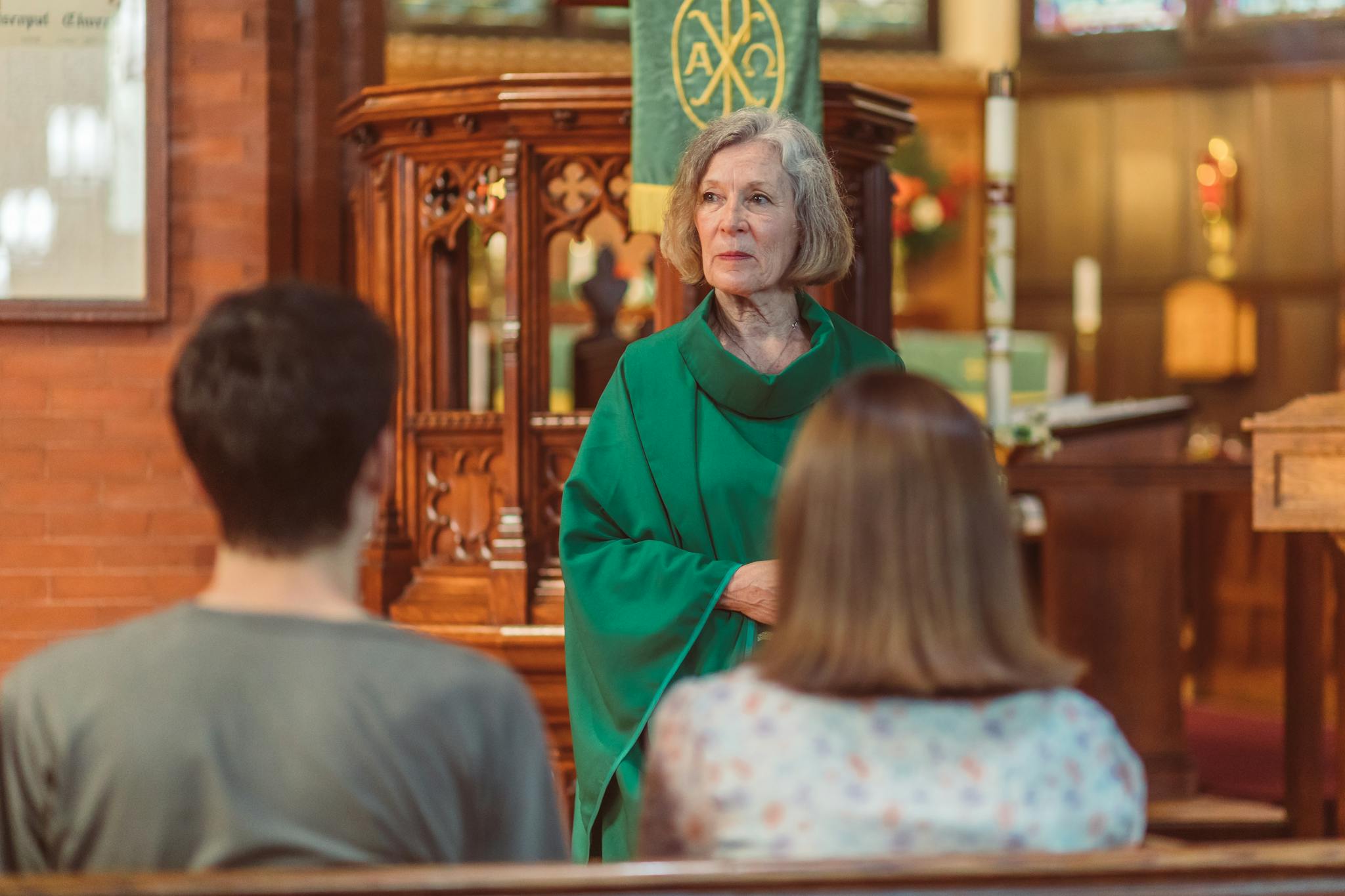 Elderly priest in green cassock leads a church service, engaging parishioners in an elegant wooden interior.